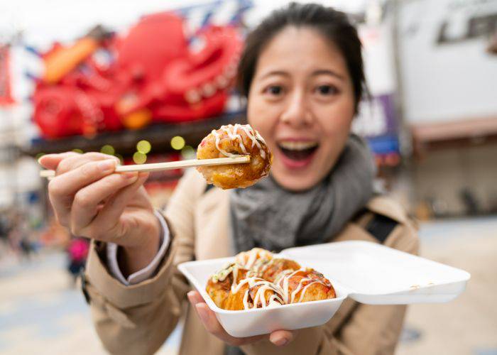 A woman holding takoyaki up to the camera, smiling.